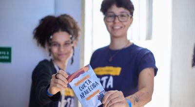Duas jovens exibem folheto do Festival Curta Aparecida, representando acolhimento e participação na programação cultural do evento.