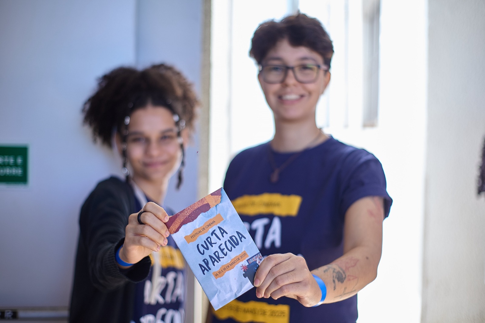 Duas jovens exibem folheto do Festival Curta Aparecida, representando acolhimento e participação na programação cultural do evento.