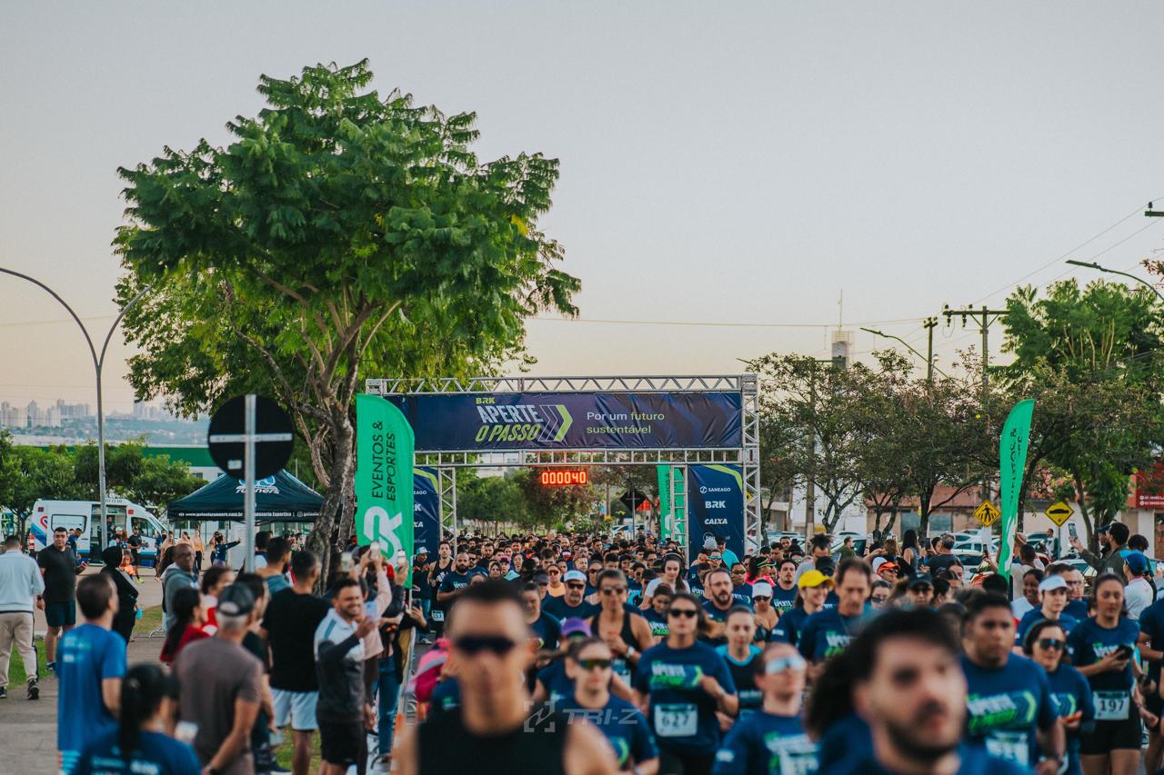 Imagem mostra a largada da Corrida BRK em Aparecida, com centenas de participantes reunidos sob pórtico do evento em clima de celebração e esporte.