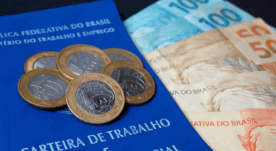 Imagem de uma carteira de trabalho, em cima contém algumas moedas e ao lado algumas cédulas de dinheiro.