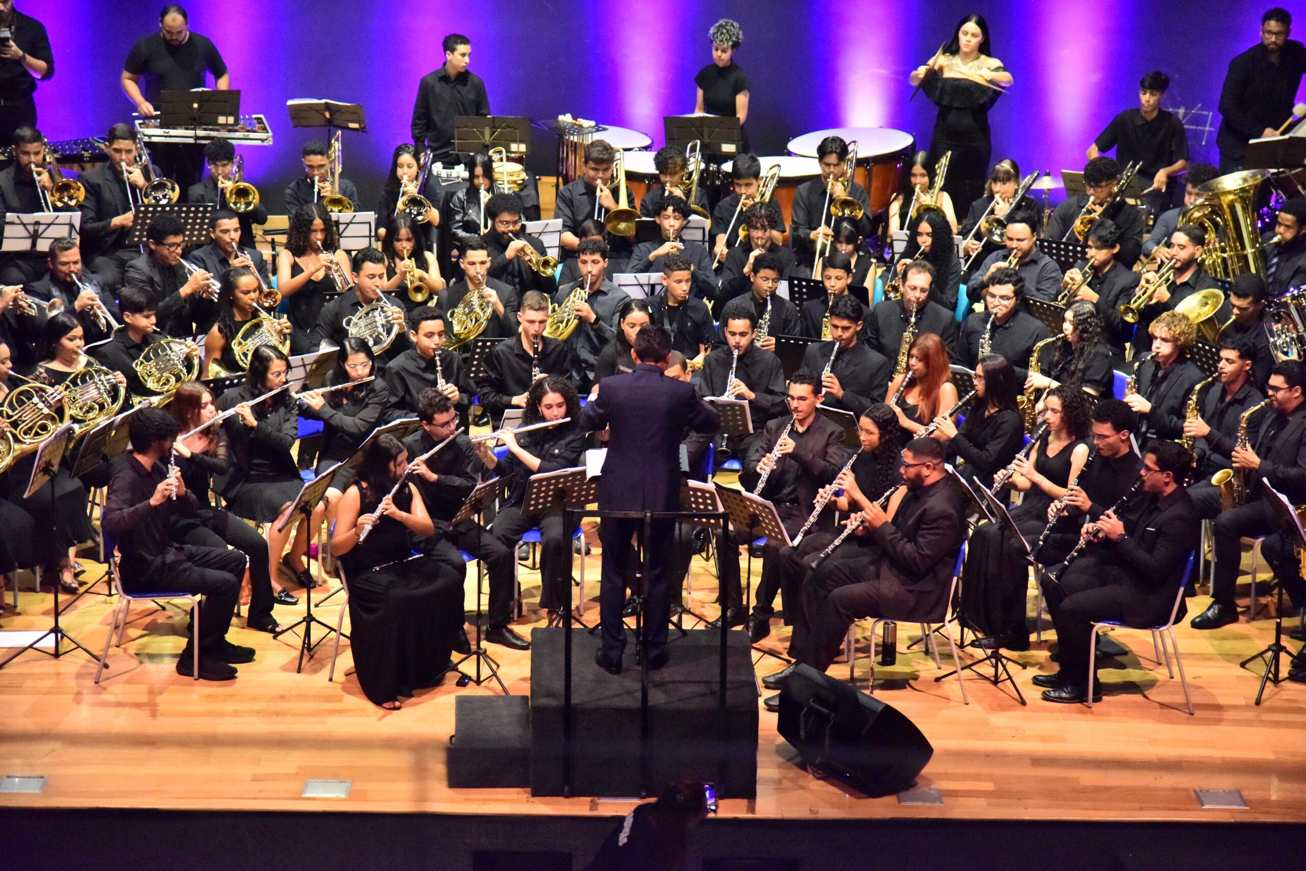 Fotografia mostra a Banda Sinfônica de Aparecida em apresentação no Anfiteatro Municipal, com cerca de 90 músicos vestidos de preto sob regência, diante de fundo iluminado em roxo.