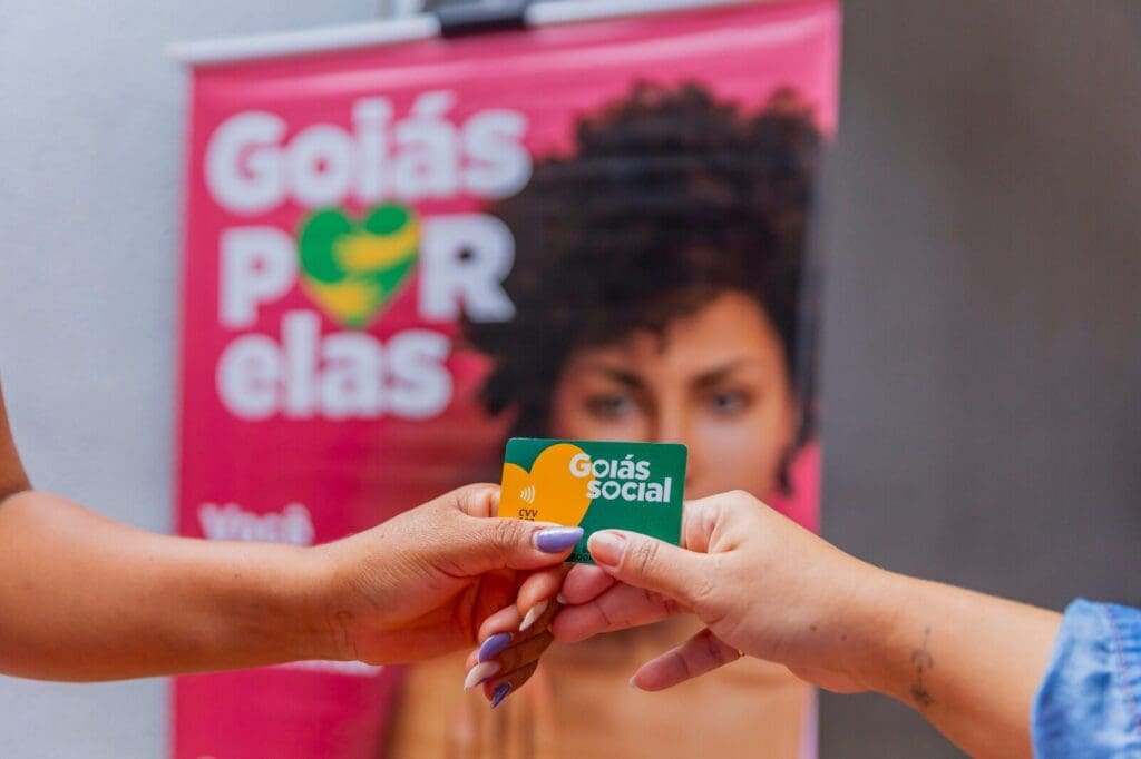 Imagem mostra entrega de cartão do programa Goiás Social, com banner do “Goiás Por Elas” ao fundo, destacando ação voltada ao apoio às mulheres.